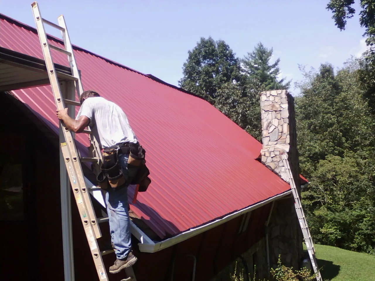 Roofer on a ladder beside a red metal roof and stone chimney — Soffit & Fascia Repair by Roofing Brothers in Collegeport